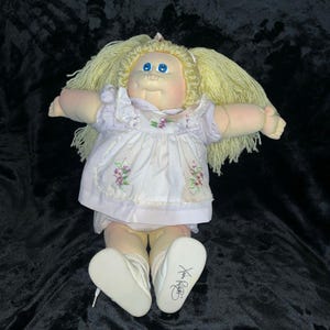 Könnte beinhalten: Eine Vintage Cabbage Patch Kid Puppe mit blonden Garnhaaren und blauen Augen. Die Puppe trägt ein weißes Kleid mit lila Blumenstickerei und weiße Schuhe. Die Füße der Puppe sind signiert.