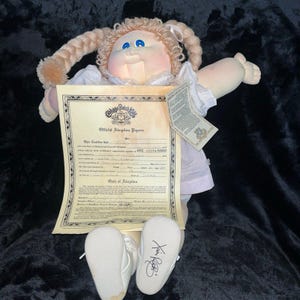 Peut inclure: Une poupée Cabbage Patch Kids avec des couettes blondes, des yeux bleus et une robe blanche. La poupée tient des papiers d'adoption et porte des chaussures blanches avec une signature sur l'une d'elles. La poupée est sur un fond noir.
