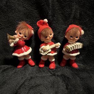 May include: Three figurines of children wearing red outfits with white trim and red hats with white pom-poms. The first child plays a gold trumpet, the second plays a white electric guitar, and the third plays a white accordion. Each has brown hair.