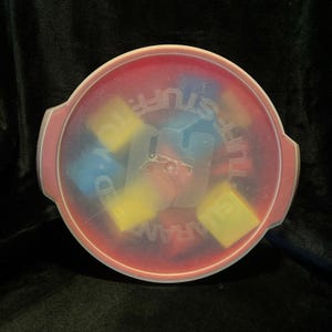 May include: A round, red plastic container with a clear lid. The lid has the words "D.A.R.A.M.A. TOY STUFF" printed on it. Inside are several yellow and blue toy blocks.