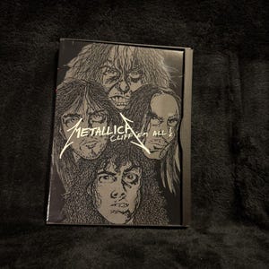 May include: A rectangular black and white VHS case featuring a graphic illustration of four faces with long hair. The text Metallica Cliff 'Em All is printed in white across the center of the cover. The design uses high contrast ink lines.