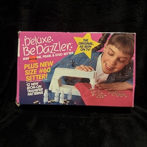 May include: A Deluxe BeDazzler rhinestone, pearl, and stud setter kit in a pink and blue box. The packaging features a white handheld tool, various setter attachments, and text including The Original, As Seen On TV and Plus New Size #60 Setter.