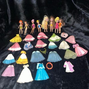 Op de afbeelding: Een verzameling miniatuurpoppen en poppenkleding. De poppen hebben verschillende haarkleuren en dragen badpakken. De kleding omvat jurken in geel, roze, blauw en wit, samen met een roze jas en een oranje ring.