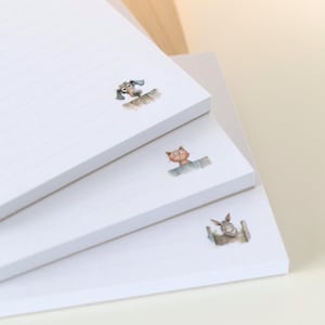 Könnte beinhalten: Drei weiße Notizblöcke, die schräg gestapelt sind, jeder mit einer anderen Aquarellillustration eines Tieres. Der obere Notizblock hat einen Hund, der mittlere eine Katze und der untere ein Kaninchen. Auf dem oberen Notizblock steht das Wort "VERGESSEN".
