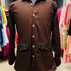 Puede incluir: Una camisa marrón con botones, dos bolsillos negros y botones decorativos. La camisa se exhibe en un maniquí. La camisa tiene cuello y mangas largas. El maniquí está sobre un soporte.