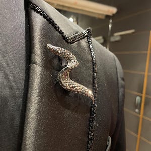 May include: A black blazer with a decorative snake brooch. The brooch is silver-toned with a textured, gem-like pattern. Black beading accents the lapel and pocket edge, adding a touch of elegance to the formal wear. The blazer is made of a shiny fabric.