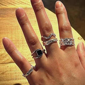 Puede incluir: Una mano con cuatro anillos de plata. Los anillos son de diferentes estilos, incluyendo un diseño de onda, un diseño de cadena, un diseño cuadrado con una piedra negra y una banda simple.