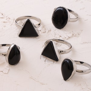 May include: Five silver rings with black gemstone settings. The rings feature different shapes, including a teardrop, oval, and triangle.