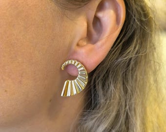 May include: Gold-colored, fan-shaped earrings. The earrings have a curved top and a series of vertical lines that create a textured, geometric design. The earrings are worn on the ear.