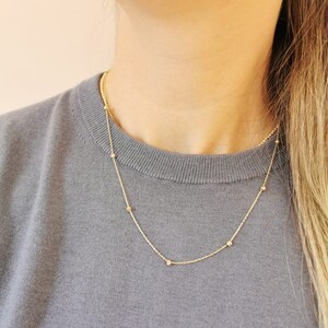 May include: A gold necklace with small, spherical beads spaced along the chain. The necklace is shown against a grey jumper, emphasising its delicate design. The necklace is a simple and elegant piece of jewellery.