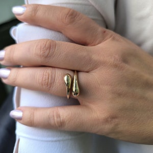 Op de afbeelding: Een gouden ring met twee traanvormige ontwerpen. De ring wordt aan een vinger gedragen.
