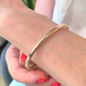 May include: A polished gold bangle bracelet with a smooth, rounded surface and a hinged clasp mechanism. The bracelet features a continuous, solid metallic finish that reflects light, creating a bright, reflective appearance on the wrist.