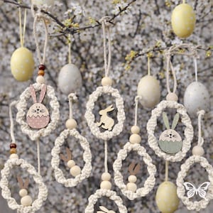 Könnte beinhalten: Osterdekorationen mit hängenden Makramee-Kränzen, Holz-Hasen- und Schmetterlingsausschnitten sowie gesprenkelten gelben und weißen Ostereiern. Die Dekorationen sind an Zweigen mit weißen Blüten aufgehängt.