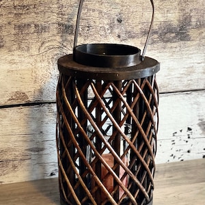 Könnte beinhalten: Eine braune Weidenkerzenlaterne mit einem Metallgriff und einem Glaseinsatz für eine Kerze. Die Laterne steht auf einer Holzoberfläche.