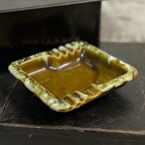 May include: A square ceramic ashtray with a brown glaze and green and white speckles. The ashtray has four indentations for cigarettes.