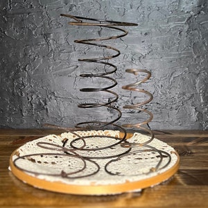 May include: A vintage metal spring coil, stacked on top of a round wooden base with a white crocheted doily. The spring coil is rusty and has a dark brown patina.