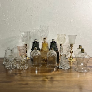 May include: A collection of 13 glass candle holders in various shapes and sizes. The candle holders are clear, frosted, and green glass. Some of the candle holders have gold accents. The candle holders are arranged on a wooden surface.