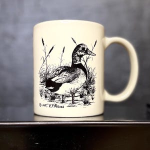 May include: Cream-colored ceramic mug featuring a black and white illustration of a duck in a marsh setting. The mug has a curved handle and the text "© 1997 K.P. Reeves" is printed on the bottom.