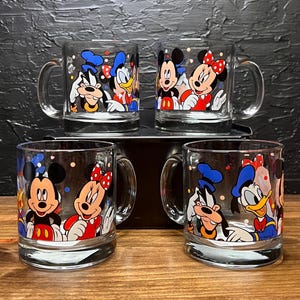 May include: Set of four clear glass mugs featuring Disney characters Mickey Mouse, Minnie Mouse, Donald Duck, and Goofy. Each mug has a handle and is decorated with colorful cartoon images. The mugs are displayed on a wooden surface.