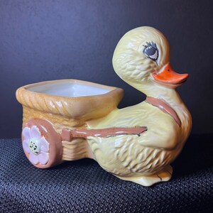 May include: A yellow ceramic duck planter with a brown and white wagon attached. The wagon has a pink flower on the wheel.