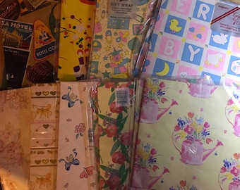 May include: Assortment of colourful gift wrap featuring various designs, including a floral pattern with pink watering cans, a baby-themed pattern with blue and pink squares, and a pattern with a clown and toys.