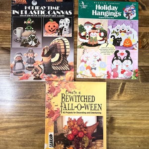 May include: Three craft books are displayed on a wooden surface. The books are titled "Holiday Time in Plastic Canvas," "Holiday Hangings," and "Create a Bewitched Fall-O-Ween." The covers feature various holiday-themed designs, including pumpkins, ghosts, and Santa.