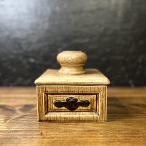 May include: A small, rectangular, beige ceramic box with a textured wood-grain appearance. It features a small drawer with a dark brown handle and a rounded, wooden-like knob on top. The box sits on a wooden surface.