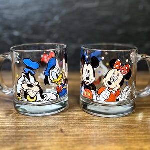 May include: Two clear glass mugs with cartoon character designs. One mug features Goofy and Donald Duck, the other Mickey and Minnie Mouse. Each mug has a handle and a silver base.