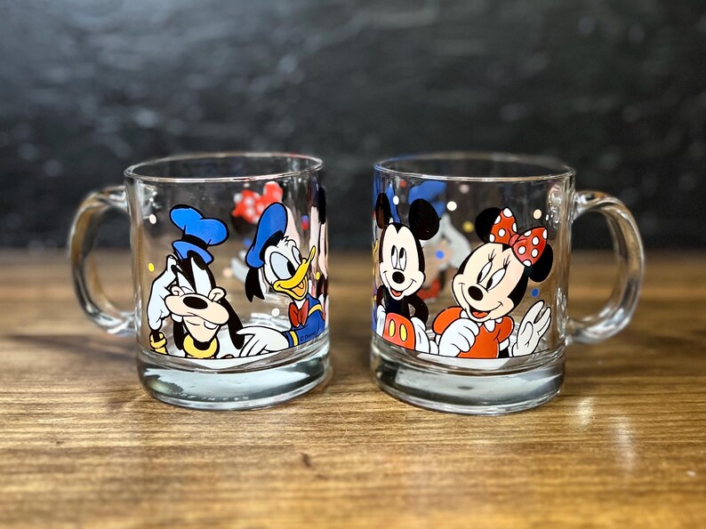 May include: Two clear glass mugs with cartoon character designs. One mug features Goofy and Donald Duck, the other Mickey and Minnie Mouse. Each mug has a handle and a silver base.