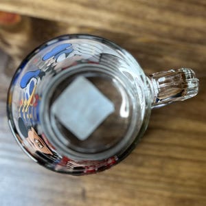 May include: Clear glass mug with a handle, featuring cartoon character designs around the rim. The mug is set against a wooden surface, viewed from above, showcasing the interior and handle details.