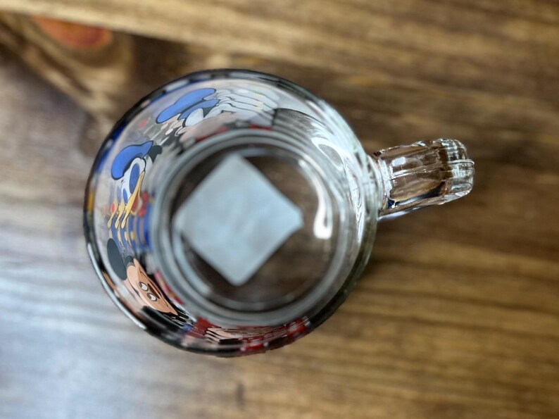 May include: Clear glass mug with a handle, featuring cartoon character designs around the rim. The mug is set against a wooden surface, viewed from above, showcasing the interior and handle details.