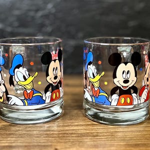 May include: Two clear drinking glasses featuring Disney characters Mickey Mouse, Donald Duck, and Goofy. The glasses have a rounded base and are decorated with colorful illustrations of the iconic cartoon characters. The Disney logo is visible on the bottom of the glasses.