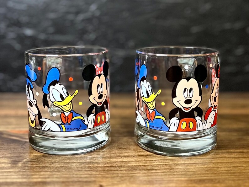 May include: Two clear drinking glasses featuring Disney characters Mickey Mouse, Donald Duck, and Goofy. The glasses have a rounded base and are decorated with colorful illustrations of the iconic cartoon characters. The Disney logo is visible on the bottom of the glasses.