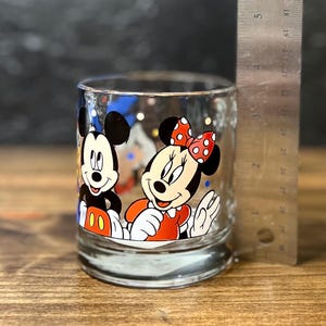 May include: Clear glass tumbler featuring Mickey Mouse and Minnie Mouse. Mickey is wearing red shorts and white gloves, while Minnie is in a red dress with a polka dot bow. The glass is approximately 3 inches tall.
