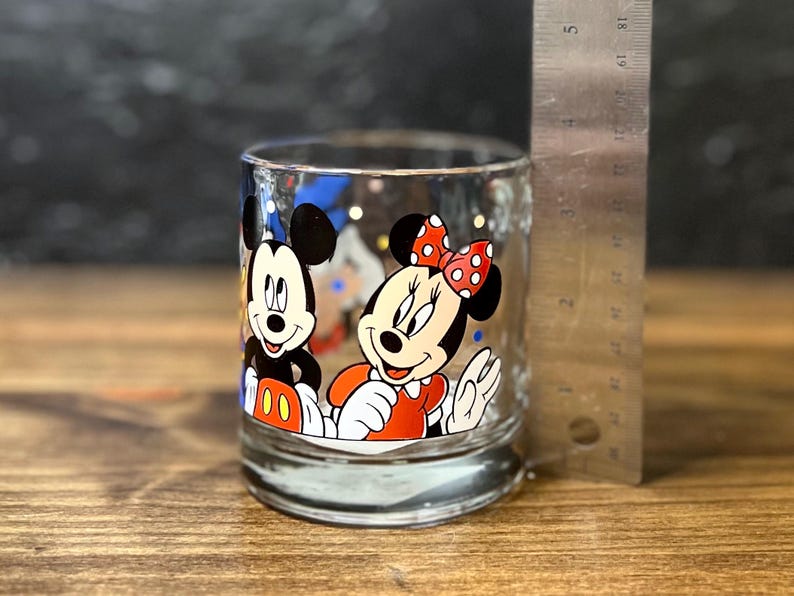 May include: Clear glass tumbler featuring Mickey Mouse and Minnie Mouse. Mickey is wearing red shorts and white gloves, while Minnie is in a red dress with a polka dot bow. The glass is approximately 3 inches tall.
