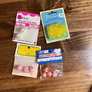 May include: Four packages of craft supplies, including white simulated pearls, yellow tri-beads, wooden doll heads, and pink wooden doll heads. The packages are labeled with the brand names "Markiseris", "Fibre-Craft", "Fibre-Craft", and "Westrim Crafts".