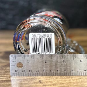 May include: Clear glass Disney mug with a handle, viewed from the bottom. The mug has a printed design and a barcode sticker. A ruler shows the mug is approximately 4 inches wide. The text on the sticker reads "NCHO HOCKING 9512 DISNEY MUG".
