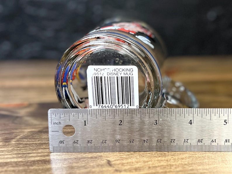 May include: Clear glass Disney mug with a handle, viewed from the bottom. The mug has a printed design and a barcode sticker. A ruler shows the mug is approximately 4 inches wide. The text on the sticker reads "NCHO HOCKING 9512 DISNEY MUG".