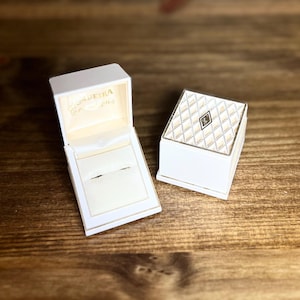 May include: Two white jewelry boxes with gold trim sit on a wooden surface. One box is open, revealing a cream-colored interior with a ring slot. The closed box has a diamond pattern on the top and a small emblem.
