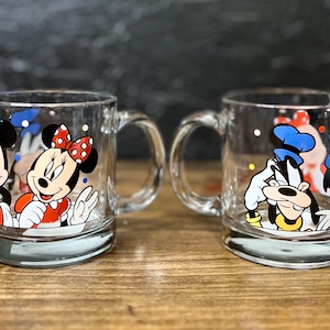 May include: Two clear glass mugs with cartoon character designs. One mug features Mickey and Minnie Mouse, the other features Goofy and Donald Duck. Each mug has a handle and a silver-colored base.