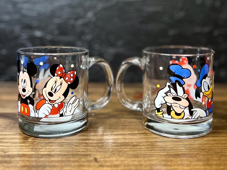 May include: Two clear glass mugs with cartoon character designs. One mug features Mickey and Minnie Mouse, the other features Goofy and Donald Duck. Each mug has a handle and a silver-colored base.