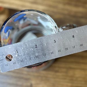 May include: A clear glass mug with a handle, partially filled, with a metal ruler laid across the top. The ruler measures up to 5 inches. The mug has colorful designs around the rim.
