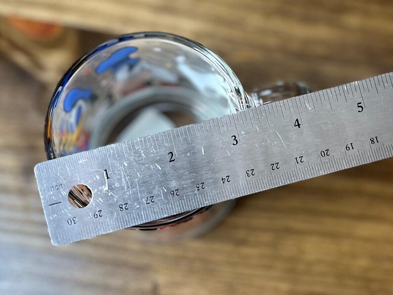 May include: A clear glass mug with a handle, partially filled, with a metal ruler laid across the top. The ruler measures up to 5 inches. The mug has colorful designs around the rim.