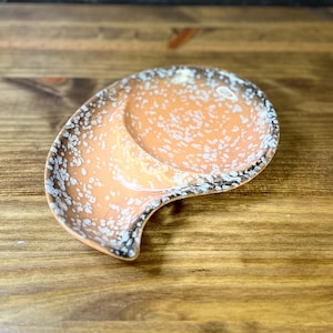 May include: A ceramic spoon rest with a speckled orange interior and a dark brown and white rim. The spoon rest has a unique, curved shape and is designed to hold a spoon or other utensil. The background is a wooden surface.