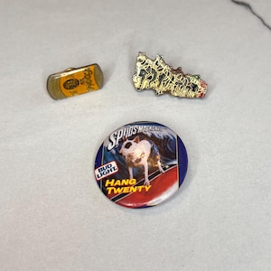 May include: Three enamel pins. One is a yellow and gold can with the word "Cooke" on it. Another is a group of dogs in a line, and the last is a round pin with a white dog on a surfboard, with the text "Spuds Mackenzie" and "Hang Twenty" on it.