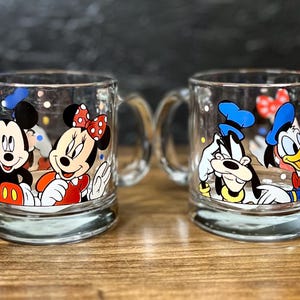 May include: Two clear glass mugs with handles, each featuring Disney characters. One mug showcases Mickey and Minnie Mouse, while the other displays Goofy and Donald Duck. The mugs have colorful character designs.