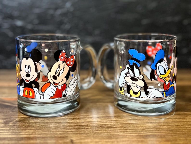 May include: Two clear glass mugs with handles, each featuring Disney characters. One mug showcases Mickey and Minnie Mouse, while the other displays Goofy and Donald Duck. The mugs have colorful character designs.