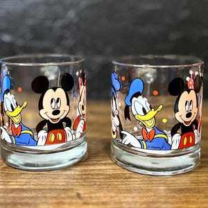 May include: Two clear glass tumblers featuring Disney characters Mickey Mouse, Donald Duck, and Minnie Mouse. The glasses have a rounded base and are decorated with colorful illustrations of the iconic cartoon characters. The Disney logo is visible on the bottom.