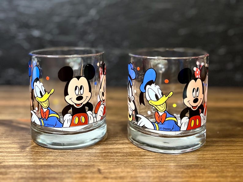 May include: Two clear glass tumblers featuring Disney characters Mickey Mouse, Donald Duck, and Minnie Mouse. The glasses have a rounded base and are decorated with colorful illustrations of the iconic cartoon characters. The Disney logo is visible on the bottom.