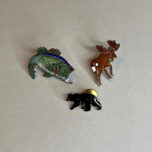 May include: Three enamel pins featuring a green and blue fish, a brown moose, and a black bear. The pins are on a white background.